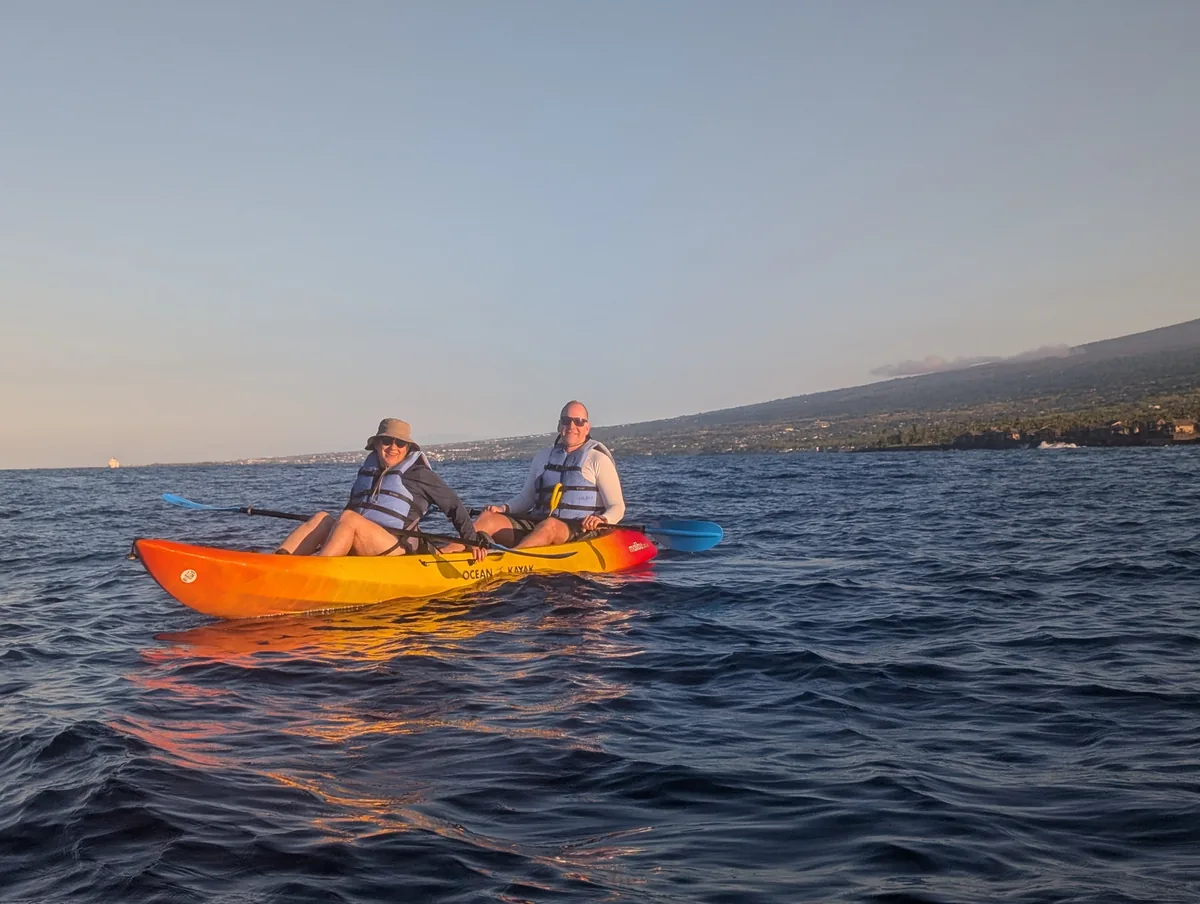 Two guests paddling an orange kayak on glassy water in the early morning light