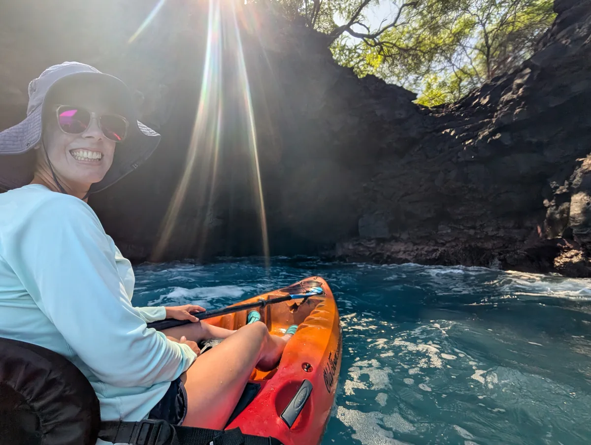 A smiling guest in her kayak as sunbeams pour into the sea cave