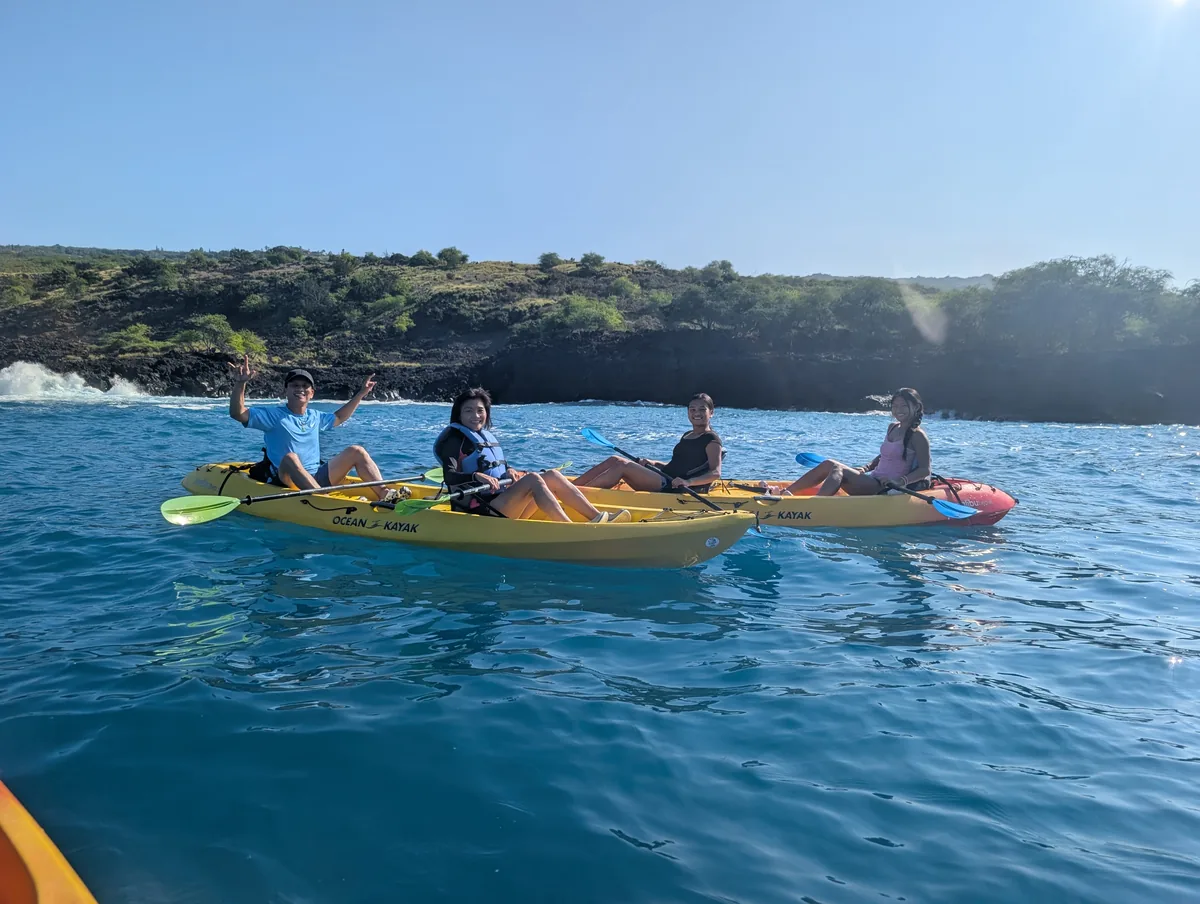 A group of smiling guests in life jackets waving from their kayaks beneath the lava coastline