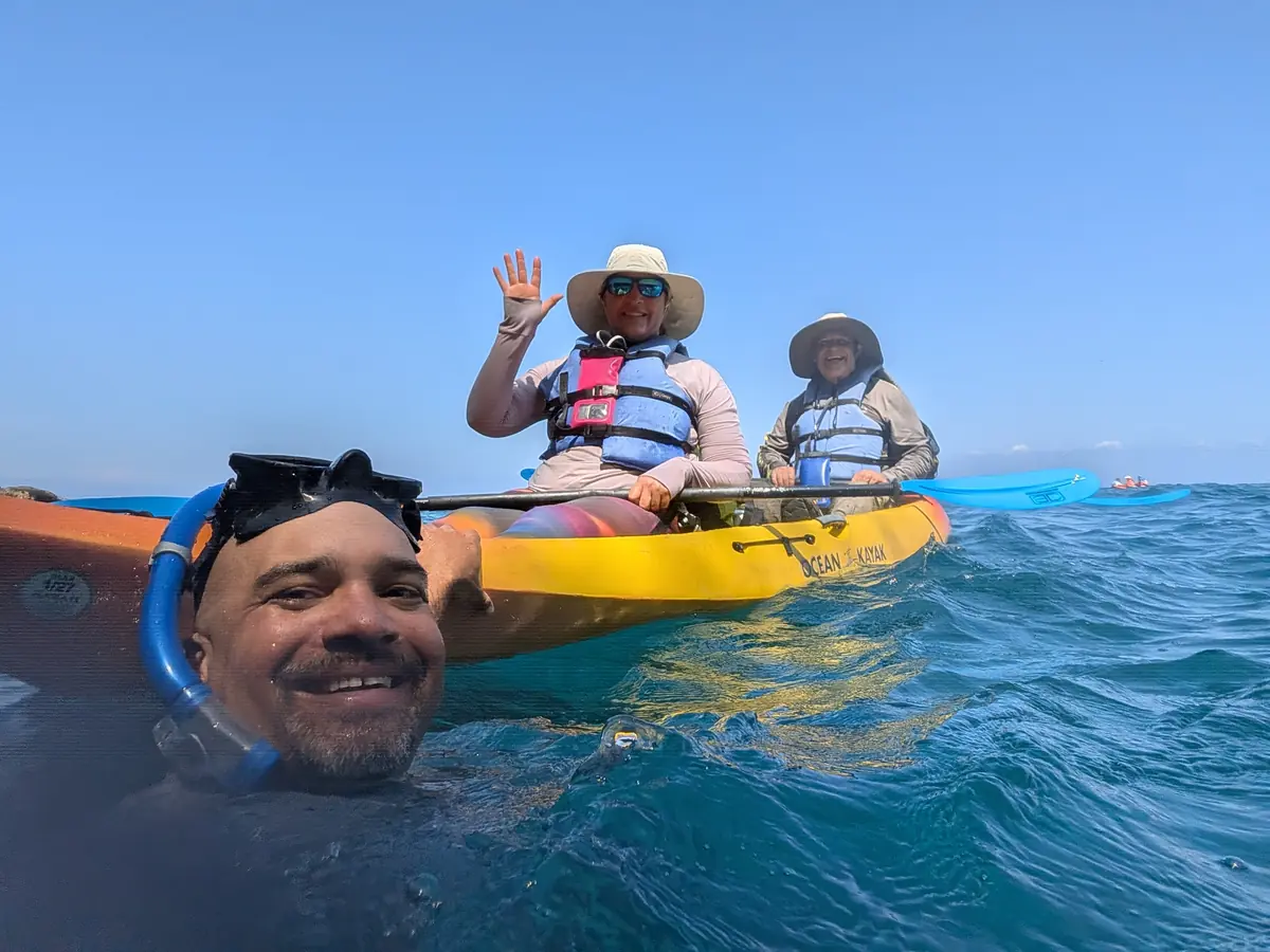 Winfield in the water beside a tandem kayak, guests in life vests waving
